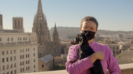 A young caucasian woman with short hair in a purple sweatshirt and black coronavirus face mask is holding dachshund dog puppy and watching in the camera. Barcelona, Spain. High-resolution jpg portrait