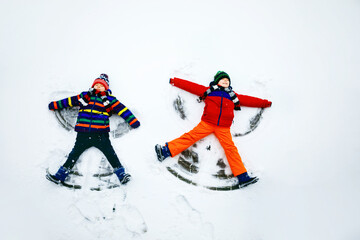 Two little siblings kid boys in colorful winter clothes making snow angel, laying down on snow. Active outdoors leisure with children in winter. Happy brothers with warm hat, gloves, winter fashion.