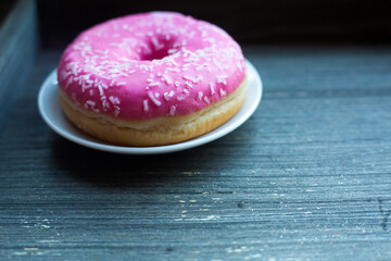 side view to bright pink donut on white round plate and aged wooden grey background. Isolated object close up. Traditional American sweets for breakfast. Copy space. White sugar decoration on top. 