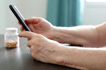 Elderly woman with smartphone sitting at the table, mobile phone in wrinkled female hands. Concept of online communication at retirement, search for disease symptoms