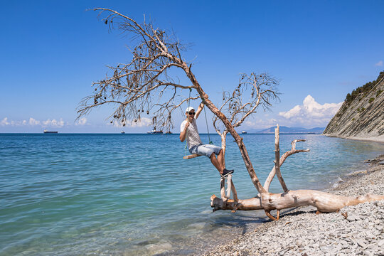 Summer landscape with a view of the sea. A guy sitting on a swing, on a fallen tree on the shore, and cargo ships on the horizon. The outskirts of the resort of Gelendzhik. Russia, Black Sea coast