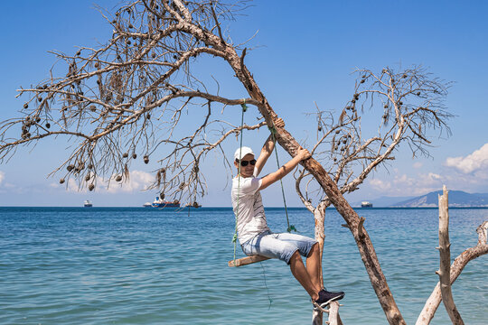 Summer landscape with a view of the sea. A guy sitting on a swing, on a fallen tree on the shore, and cargo ships on the horizon. The outskirts of the resort of Gelendzhik. Russia, Black Sea coast