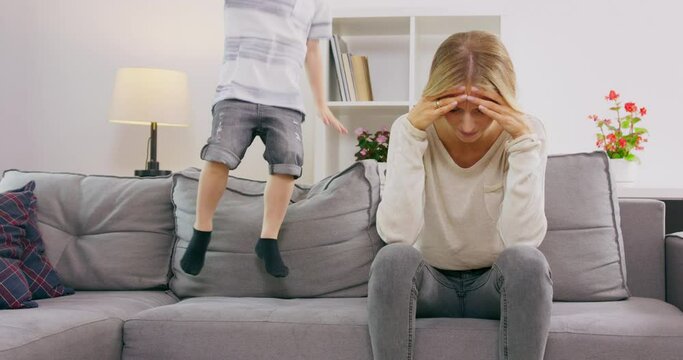 Tired Mother Covering Ears Having Headache While son Jumping On Couch At Home. Parenting Hyperactive Child.