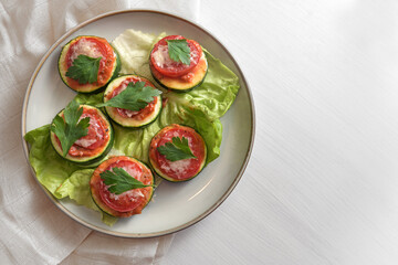 Mini pizza snack from oven baked zucchini slices, tomato, parmesan cheese and parsley garnish on a plate with lettuce leaves, white painted table with copy space, high angle view from above