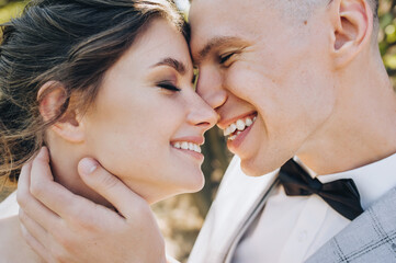 Close-up faces of a groom in a gray suit, a black bow tie, a boutonniere and a beautiful brunette bride in a white dress with curly hair on a nature background.