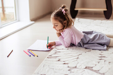 a child girl sits on the floor and draws with markers in an album. Games at home on the carpet. The interior of the apartment