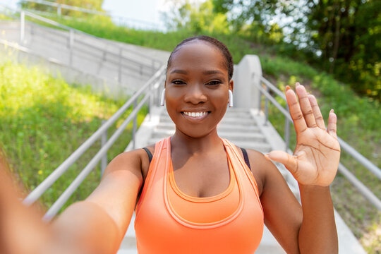 fitness, sport and technology concept - happy smiling young african american woman with wireless earphones listening to music, taking selfie and waving hand outdoors