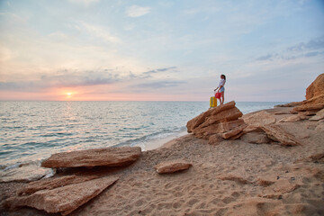 Woman with suitcase on rock watching nature sunset. Summer vacation, relaxation time and tourism concept. Copy space