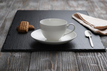 White empty cup of cappuccino, small spoon, biscuits on a black textured stone plate, wooden textured table.