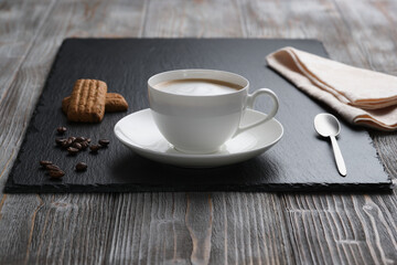 White cup of cappuccino, small spoon, cookies on a black textured stone plate, wooden textured table. White foam. Coffee beans on the table.