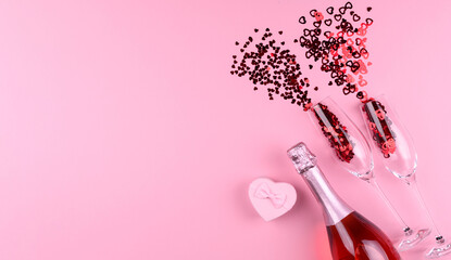 Two glasses filled with heart sequins with a bottle of champagne and a Valentine's Day gift on a pink background