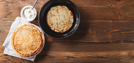 Traditional Russian Crepes Blini stacked in plate and pancake in cast-iron frying pan on wooden table. Maslenitsa Russian festival meal. Russian kitchen. Top view. Space for text. Long wide banner