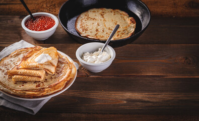 Traditional Russian Crepes Blini stacked in plate and cast-iron frying pan with red caviar, fresh sour cream on dark wooden table. Russian festival meal Maslenitsa or Shrovetide. Space for text