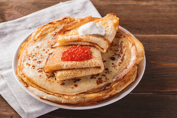 Traditional Russian Crepes Blini stacked in a plate with red caviar, fresh sour creamon dark wooden...