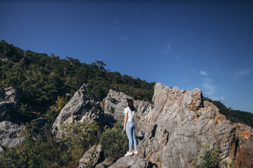 woman traveler in blue jeans and a white t-shirt with dark hair stands on a stone and looks at a rocky forest landscape