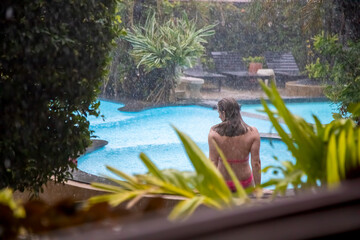 a girl in a bikini sits on the side of an outdoor pool in a tropical garden in the rain