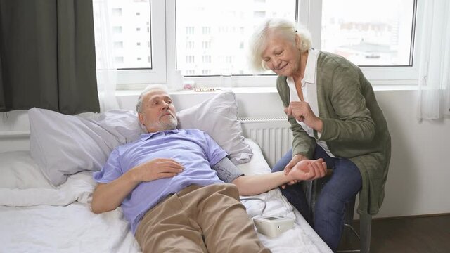 caring elderly woman measures her beloved husband's blood pressure while lying on a bed in a room.