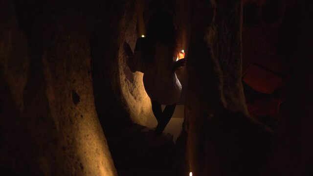 Long haired woman in white blouse with flashlight on smartphone goes down steps in handmade cave in old Cappadocia park backside view.