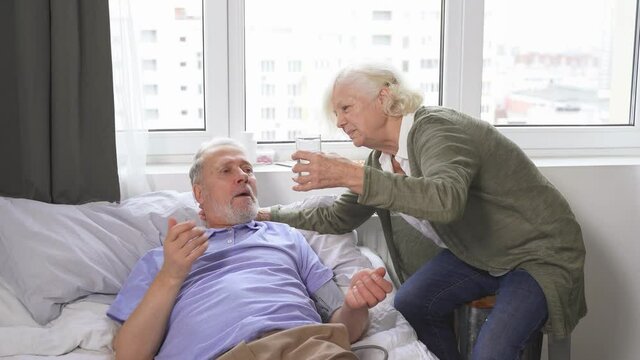 an elderly woman takes care of her husband by measuring blood pressure the wife gives her husband a pill and a glass of water, the husband lies on the bed at home. The wife's concern for her husband.