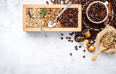 Green and brown decaf unroasted and dark roasted coffee beans in wooden box with scoops setup on white concrete background.