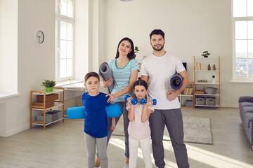 Fototapeta premium Active family sports workout in lockdown at home concept: Portrait of happy fit young couple with healthy little children standing together, holding exercise mats and dumbbells and looking at camera
