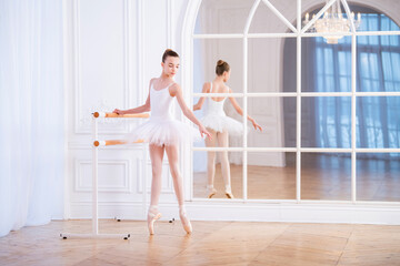 young ballerina stands on pointe shoes at a ballet barre in beautiful white hall in front of a mirror. © Maria Moroz