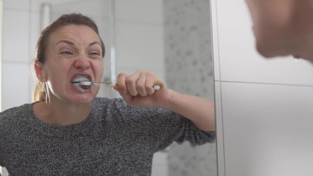 A woman aggressively brushes her teeth in the morning. Reflection in the mirror woman presses heavily on her teeth with a toothbrush.