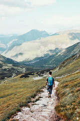 Young man with backpack hiking in a mountains, actively spending summer vacation. Rear view of teenager walking up on a hill along mountain path