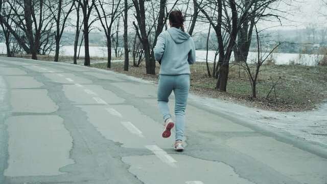 Fit woman in gray sportswear running on asphalt road in slow motion, frozen lake on background. Following shot of female athlete training outside in winter. Concept of fitness
