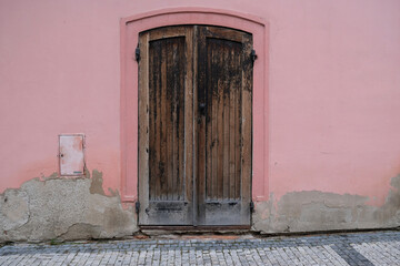 Vintage wooden painted door of old building. Shabby door boards. Old wall made of pink plaster and gray putty stains on the wall. Smooth surface. Cracks. Old peeling plaster wall, crumbles.