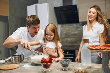 A full multicultural family with an adorable daughter gathered in a modern kitchen, preparing pancakes together. Making cake mix, pouring milk, socializing pleasure and culinary hobby