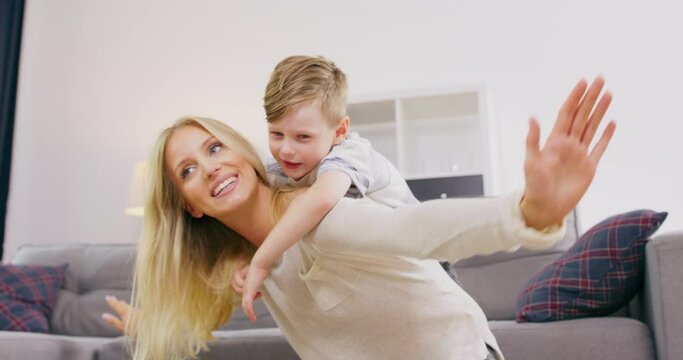 Happy beautiful family healthy young mom lifting cute little child boy up playing plane on carpet at home, funny kid boy flying in mothers arms practice acro pair yoga bonding having fun together.