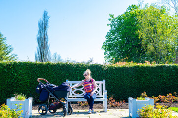 Young mother with a baby sling carrier and carriage for her newborn sitting on a park bench