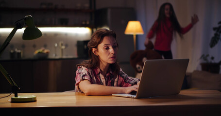 Young tired mother working at home with laptop with little daughter jumping on couch on background