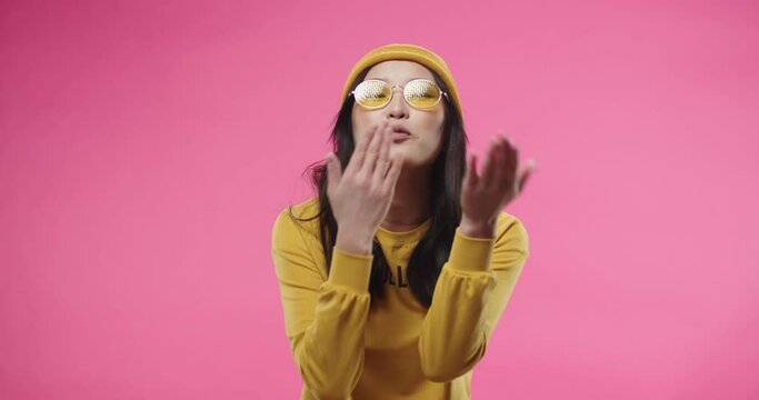 Portrait of young cheerful Asian beautiful woman in yellow hat and stylish glasses sending air kisses and making heart gesture, feeling thankful and happy while standing isolated on pink background.