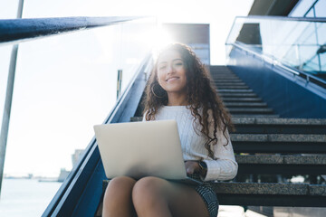 Portrait of happy Spanish millennial copywriter sitting at urban stairs with modern laptop computer...