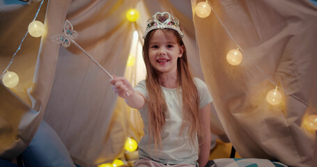 Portrait of adorable little girl wearing crown and holding magic wand sitting in teepee tent at home