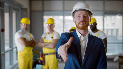 Contractor in hardhat stretching hand for handshake at camera with builders on background