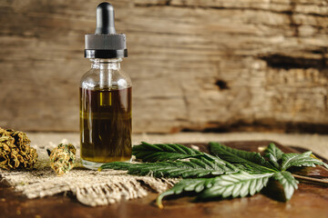 a vial with a dropper of cannabis concentrate cbd stands next to dried marijuana buds and a cannabis leaf on a rusty wooden and sackcloth background. copy space