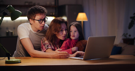 Happy family with kid sitting at table using laptop at home in evening