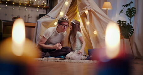 Father and daughter wearing crowns playing in toy wigwam at home