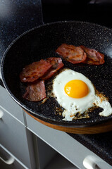 Vertical image of pan and fried egg, bacon on it, black kitchen table.Homemade breakfast