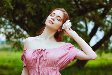 Outdoor portrait of young redhead woman in field of green grass with pink peony. Woman's Day. Female spring, summer fashion concept.