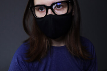 Young woman wearing stylish fashionable thick-rimmed glasses and medical black fabric mask. Dark background. 