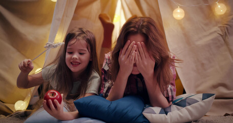 Mother and daughter lying in tent and playing together in evening at home