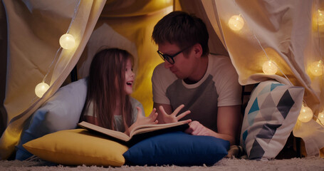 Happy father and daughter lying on floor in teepee and reading book together at home © TommyStockProject