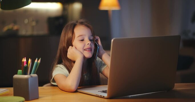 Little girl talking on cellphone and watching cartoon on laptop sitting at table in dark living room