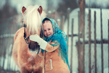 Russian style. Beautiful little girl in blue pavloposad shawl is feeding pony in winter in Russia....
