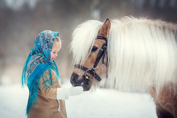 Russian style. Beautiful little girl in blue pavloposad shawl is feeding pony in winter in Russia. Image with selective focus and toning