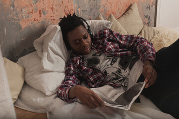 a young african american man lies on the bed wearing headphones and reads a magazine